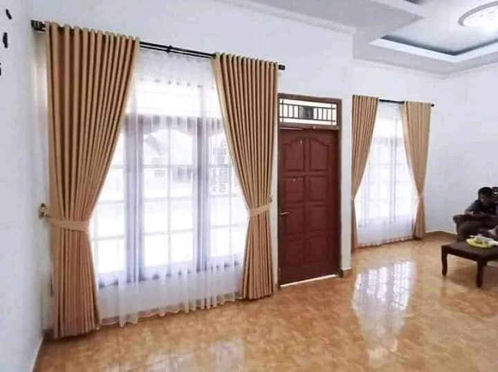 Rel pipa hitam panjang dengan beberapa bracket menopang gorden krem dan vitrase putih di sepanjang jendela ruang tamu