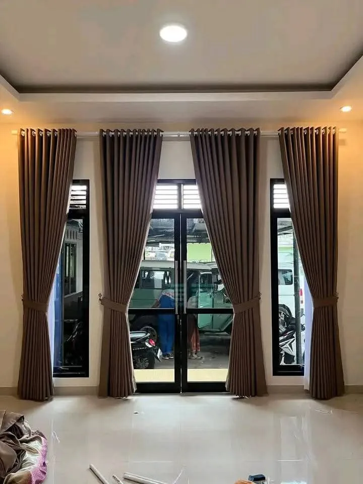 Empat panel gorden cokelat tua menutupi pintu kaca berbingkai hitam dan dua jendela di sampingnya pada ruangan berplafon terang