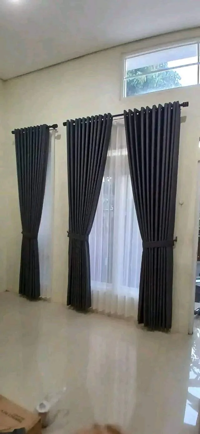 Tiga panel gorden smokring abu tua pada pipa hitam dengan vitrase putih di belakangnya, terpasang di ruang berlantai keramik