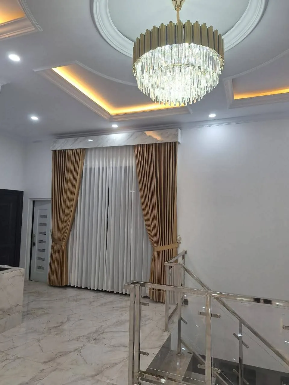 Gorden kain warna emas dengan vitrase putih dan kanopi bermotif marmer di ruangan berplafon tinggi dengan lampu gantung kristal