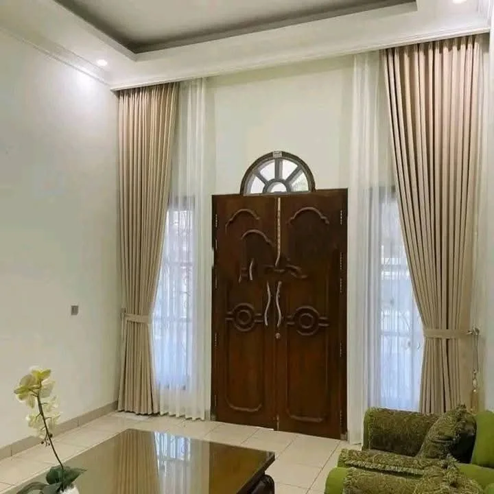 Vitrase putih dipasang bersama gorden krem di samping pintu kayu ukir, ruang tamu tetap terang di siang hari